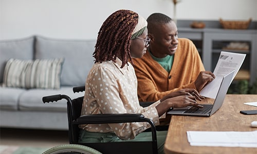 Woman in wheelchair types on laptop while reviewing paperwork with man.